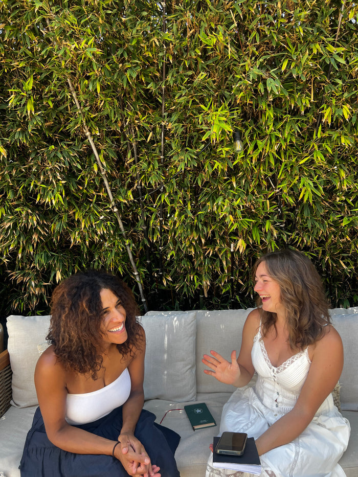 Two women sitting on a couch outdoors with a green bush in the background