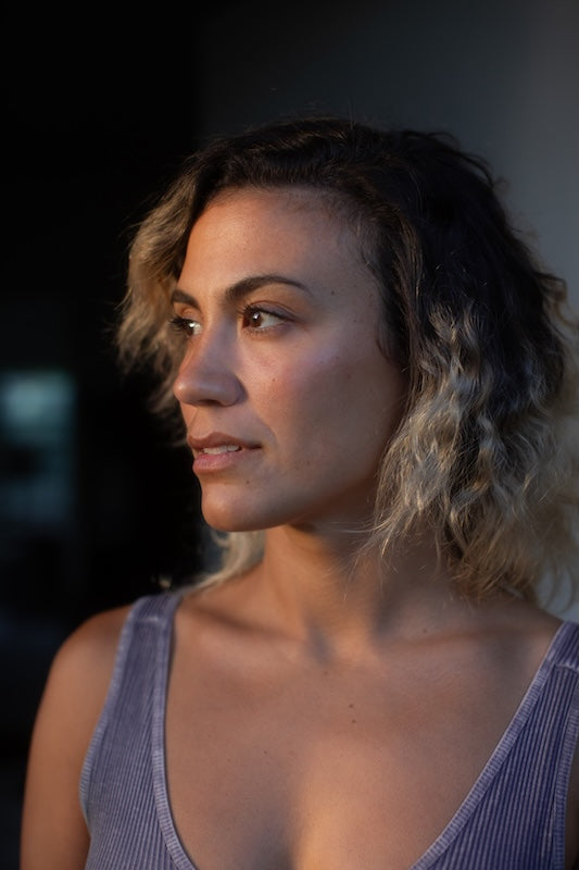 A close-up portrait of a woman with wavy shoulder-length hair, wearing a ribbed lavender tank top, captured in warm side lighting against a dark background.