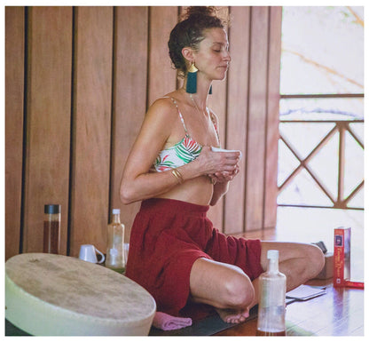 Woman sitting on a mat with a cup, surrounded by yoga props and books in a wooden room.