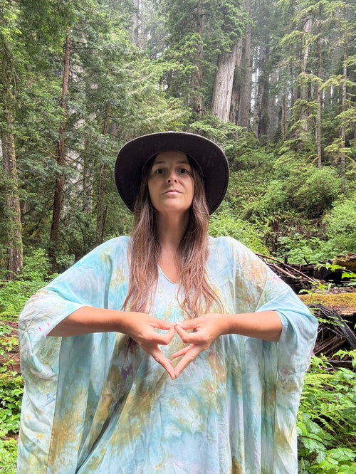 A woman wearing a wide-brimmed black hat and a light blue tie-dye kaftan poncho stands in a lush green forest, forming a heart shape with her hands.
