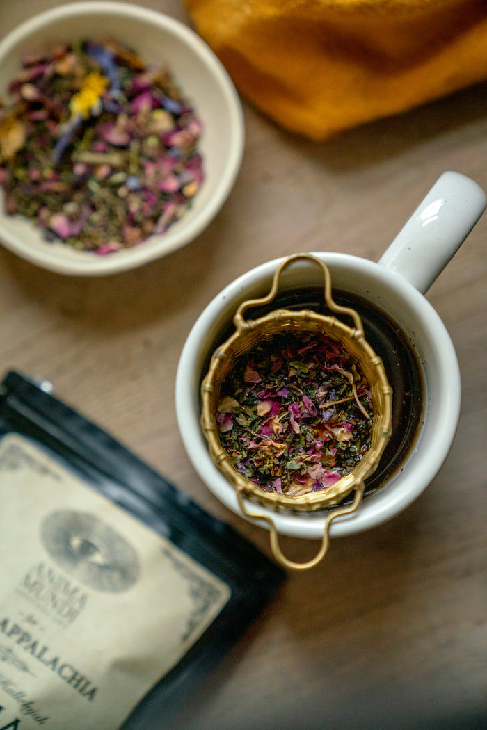 Tea cup with a gold tea infuser filled with colorful tea leaves, next to a bowl of more tea leaves and a package on a wooden surface.
