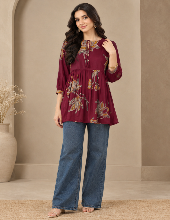 A woman modeling a three-quarter sleeve tunic top in deep maroon with a yellow and gold floral print, paired with blue jeans, in a warm beige interior setting.