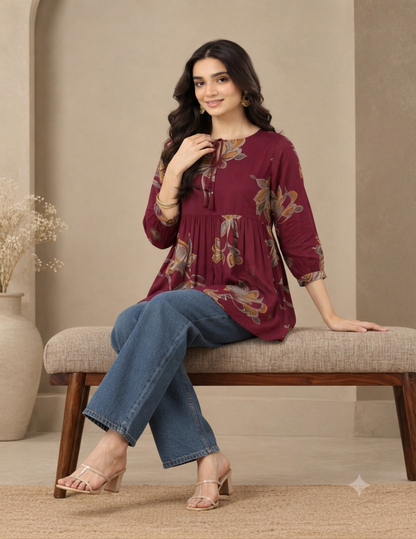 A woman models a burgundy tunic top with a brown and gold floral print, paired with blue jeans and beige sandals, sitting on a beige bench in a neutral indoor setting.