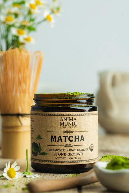 Jar of Anima Mundi Matcha on a wooden surface with flowers and a whisk in the background
