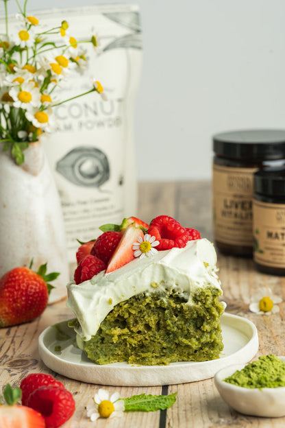 Green cake with white frosting and strawberries on a wooden table with coconut cream powder and other ingredients in the background.